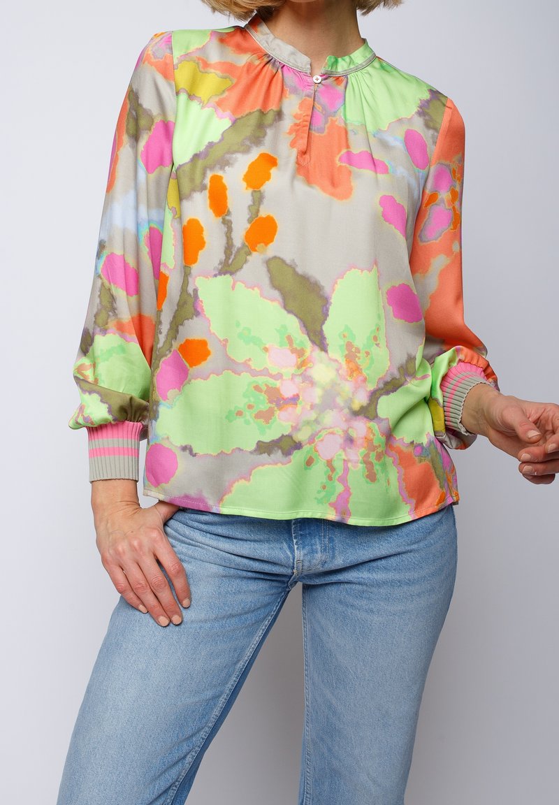 Femme portant un chemisier à manches longues coloré à motifs floraux rose, orange, vert et beige, associé à un jean en denim bleu clair.