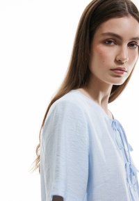 Blusa azzurro chiaro con finitura texturizzata, maniche corte e scollatura rotonda. Presenta una chiusura con fiocco nella parte anteriore.