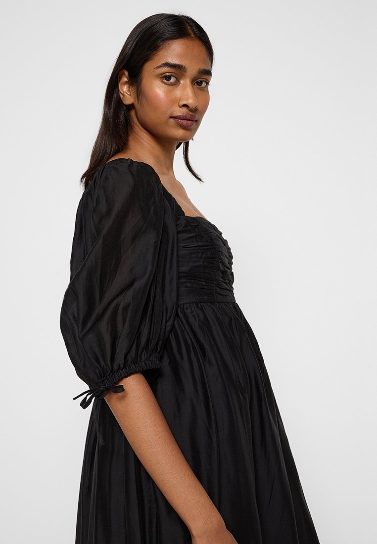 Robe noire en tissu léger, avec un corsage froncé, une jupe pleine et des manches bouffantes avec des poignets élastiques.