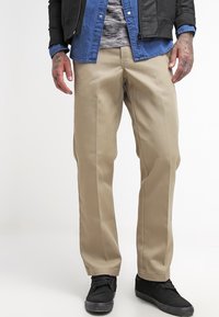 Beige chinos med lige ben-design, der har en glat tekstur og en frontfold. Parret med en sort bomberjakke og en blå skjorte.