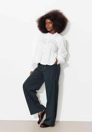 Femme aux cheveux bouclés volumineux portant un chemisier blanc avec des détails en dentelle, un pantalon large à fines rayures noires et des mocassins marron, debout contre un mur blanc.