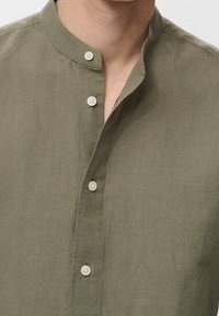 Olivgrüne Leinenbluse mit Stehkragen, dreiknöpfiger Knopfleiste mit runden weißen Knöpfen und einer weichen, strukturierten Oberfläche.