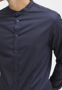 Camisa azul marino de botones con cuello mao, textura suave, diseño ajustado y detalles de costura sutil a lo largo de los botones.