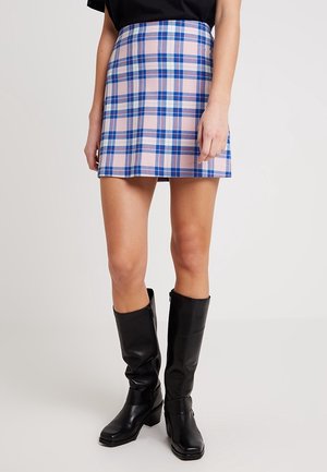 Femme portant une mini-jupe à carreaux rose et bleue avec des bottes en cuir noires montantes jusqu'aux genoux, debout devant un fond clair uni.