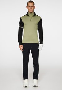 Idrottslångärmad topp i olivgrön och svart, med dragkedja i halsen och randig accent. Matchad med svarta byxor och vita sneakers.