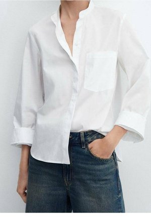 Osoba ubrana w luźną białą koszulę z długim rękawem i kieszenią na piersi oraz ciemnoniebieskie dżinsy, z ręką w kieszeni dżinsów, na jednolitym tle.