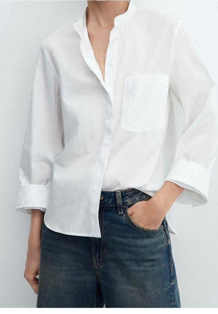 Personne portant une chemise blanche ample à manches longues avec une poche poitrine et un jean bleu foncé, main dans la poche du jean, sur un fond uni.