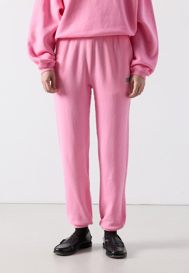 Cilvēks, kurš valkā vaļīgas rozā sporta bikses un atbilstošu džemperi, melnās zeķes un melnus loaferus, stāv uz gaišas grīdas.