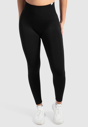 Zwarte high-waisted leggings gemaakt van rekbare stof, met een gladde textuur en een brede tailleband. G gedragen met witte sportschoenen.