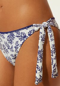 Vit bikiniunderdel med blå korallmönster, med en texturerad tyg och detalj med knytning på sidan.