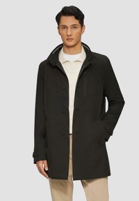 Cappotto impermeabile nero con tasca sul petto con zip, chiusura a bottone e polsini regolabili. Indossato su una polo crema e pantaloni beige.