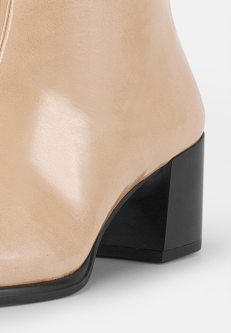 Beige enkellaarsje met een glanzende afwerking, voorzien van een vierkante zwarte hak en een gladde leren textuur. Eenvoudig ontwerp zonder zichtbare patronen.