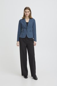 Naine seisab ja vaatab otse, kandes sinist jopet, musta särk üle, mustad laia säärega püksid ja mustad kingad heledal ühtlasel taustal.