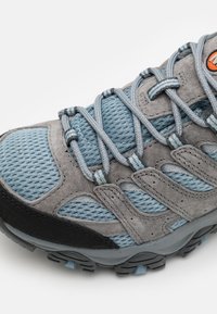 Grå og blå sneakers med mesh- og semsket skinn-materialer, teksturert overflate, lagdelt design, og flate lisser med en oransje logo-akse.