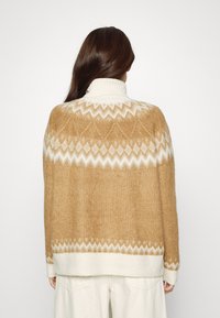 Rostfärgad tröja med ett vitt geometriskt mönster runt axlarna och en hög ribbad krage; mjuka, strukturerade stickade material.