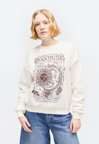 Jeune personne portant un sweat crème avec un motif graphique marron représentant le soleil, la lune, une fleur et le texte « Mystical Moonrise » ainsi qu'un jean bleu sur fond uni.