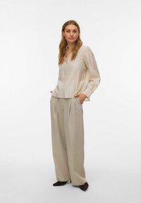 Gaiši bēša blūze ar garām piedurknēm, V veida izgriezumu un smalku tekstūru, kombinēta ar augsta vidukļa, platu kāju biksēm līdzīgā tonī.