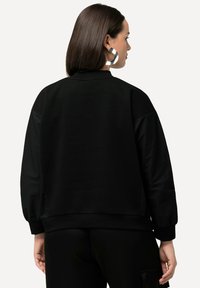 Sudadera negra con un tejido suave, cintura ajustada y mangas abullonadas. Presenta paneles contrastantes en los hombros y la espalda.