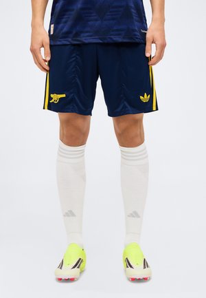 Marineblauwe voetbalshorts met gele Arsenal-kanon- en Adidas-logo’s, gedragen met witte kniekousen en neongele voetbalschoenen.