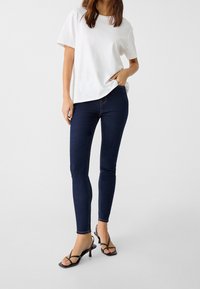 White cotton t-shirt paired with dark blue skinny jeans featuring light stitching. Worn with black heeled sandals, showcasing a minimalist design.