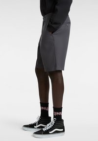 Graue Shorts mit flachem Bund, Seitentaschen und Gürtelschlaufen. Kombiniert mit schwarzen High-Top-Sneakern und gemusterten schwarzen Socken.