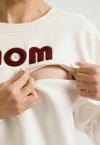 Witte sweatshirt met bordeauxrood gestructureerde "mom" letters, die wordt opgetrokken om de huid eronder te onthullen. Zachte stof en relaxte pasvorm.