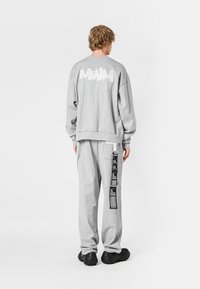 Sudadera gris con gráfico texturizado en blanco en la parte posterior, combinada con pantalones de chándal gris a juego que presentan una etiqueta impresa en el lateral. Calzado negro.