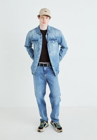 Lichtblauwe denimjas en jeans, zwart T-shirt, zwarte riem en multi-kleurige sneakers. Hoed is beige met een logo. Casual fit.