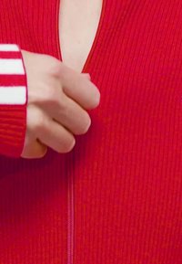 Red ribbed cardigan with a zipper and a deep neckline, featuring white stripes on the sleeve. The fabric appears textured and form-fitting.