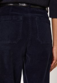Pantaloni in corduroy blu navy con tasche posteriori a superficie texturizzata. Una cintura nera completa il girovita. Design semplice e funzionale.