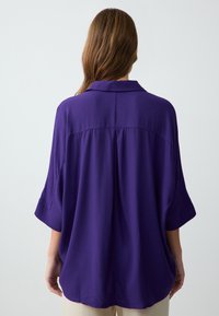 Chemise violette ample avec un col, manches larges courtes et ourlet droit. Texture douce sans motifs ni éléments métalliques visibles.