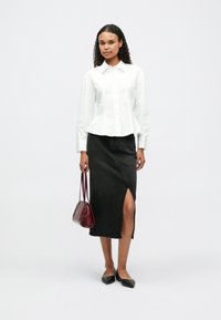 Chemise blanche à manches longues avec une taille ajustée, jupe en denim noir avec une fente sur le côté, chaussures noires à plat, et un petit sac à main bordeaux.