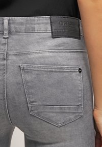Jean gris de denim con un bolsillo y una parche de cuero negro en la cinturilla. La tela tiene una textura suave con detalles de costura sutiles.