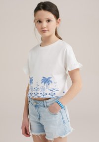 Camiseta blanca corta con diseño de palmera y playa bordado en azul, con mangas con volantes. Combinada con pantalones cortos de mezclilla claros.