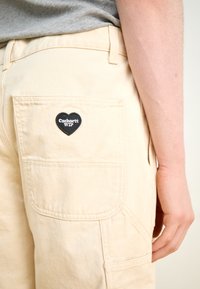 Beige bomullsbukser med sidelomme og en svart hjerteformet lapp merket "Carhartt WIP" sydd på baklommen. Glatt tekstur.