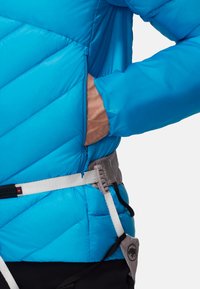 Giacca isolante blu con motivo a diamante, tasca visibile e polsini elasticizzati, indossata sopra un imbrago da arrampicata. Il materiale sembra liscio e leggero.