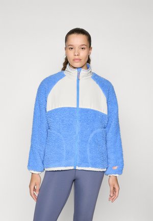Blå fuzzy fleecejakke med lys grå overdel, høy krage, frontglidelås og sidelommer. Har oransje detaljer og en teksturert overflate.