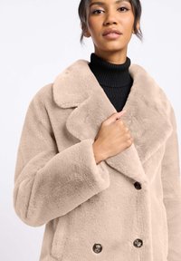 Beige nepbontjas met brede revers, dubbele knoopsluiting en lange mouwen. De textuur lijkt zacht en pluizig.