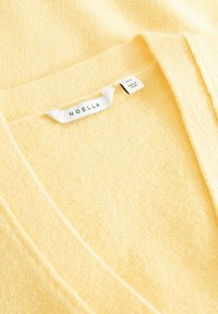 Měkký žlutý pletený svetr s výstřihem do V a bílou značkou uvnitř s nápisem „Noella“ a menší visačkou s nápisem „Vyrobeno v Číně.“