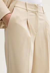 Pantalones tailored en beige claro, confeccionados con un tejido suave. Presentan un diseño de frente plano, bolsillos laterales y un corte recto.