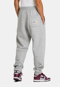Graue Sweatpants mit lockerer Passform, ausgestattet mit Seitentaschen und einer Gesäßtasche mit einem Logo-Patch. Ergänzt durch kastanienbraune Sneakers.