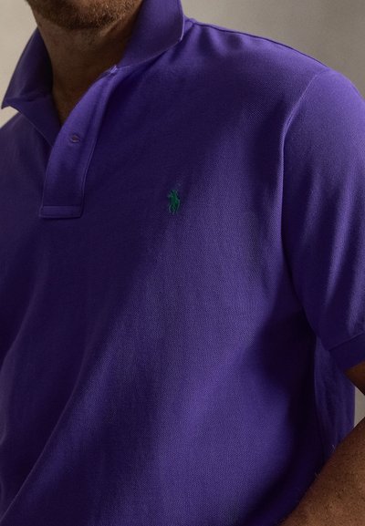 Μωβ πόλο μπλουζάκι με κοντά μανίκια, κατασκευασμένο από υφασμα με υφή. Διαθέτει ένα μικρό πράσινο έμβλημα στο αριστερό στήθος. Κλασικό στυλ γιακά.