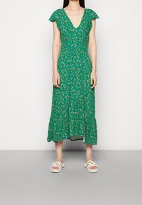 Grünes Blumen-Midi-Kleid mit kurzen Schmetterlingsärmeln, V-Ausschnitt und Rüsche am Saum. Getragen mit weißen geflochtenen Sandalen.