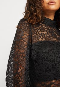 Haut en dentelle noire avec motifs floraux, manches transparentes et col montant. Le tissu est délicat et texturé, ajoutant un intérêt visuel.
