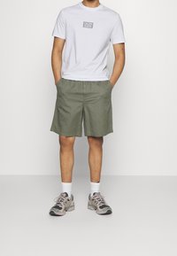 Camiseta blanca de algodón con un pequeño logo rectangular gris, combinada con pantalones cortos verdes oliva y zapatillas ligeras con acentos grises y rosas.