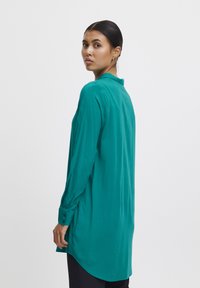 Long-sleeve teal blouse with a collar, buttoned cuffs, and a rounded hemline, made from a smooth fabric with a slightly loose fit.