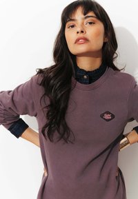 Sweatshirt violet avec un col rond et une patte en forme de diamant portant l'inscription "Sister Club". Il présente un col en denim et des manches texturées.
