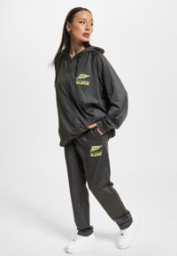 Sivá mikina na zips a zodpovedajúce tepláky vyrobené z mäkkej tkaniny, s jasne žltými logovými akcentmi a uvoľneným strihom. Biely tenisky dotvárajú outfit.