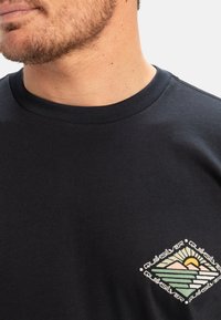 Svart bomullstrøye med et flerfarget grafisk trykk på brystet som viser fjell og en sol, med tekst som rammer inn designet.