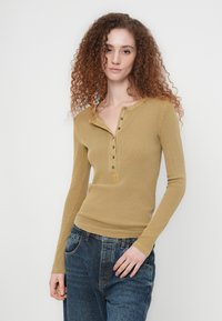 Μπλούζα henley με ριμπ δομή, μουσταρδί χρώμα, μακριά μανίκια, στρογγυλή λαιμόκοψη, πέντε κουμπιά και διακοσμητικά ράμματα. Συνδυάζεται με φαρδιά μπλε τζιν παντελόνια.